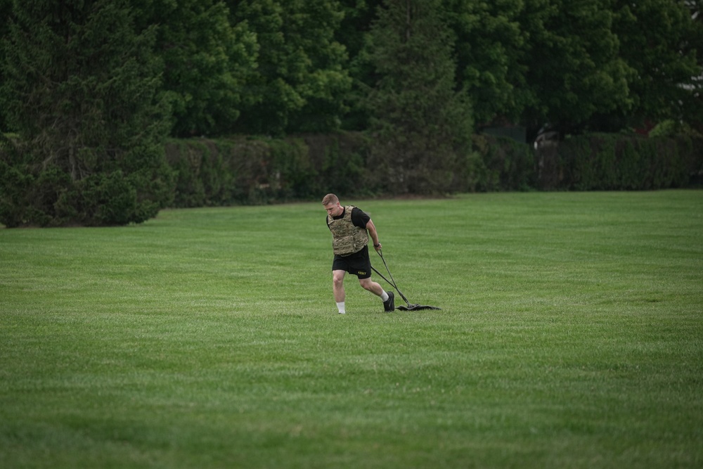 A male athlete is performing a resistance sled drag exercise on a well-maintained green grass field. He is wearing military-style workout attire including a camouflage plate carrier vest, black shorts with 'ARMY' text, and is pulling a weighted sled using straps. The setting appears to be an open field surrounded by dense green trees, suggesting an outdoor military or athletic training session.