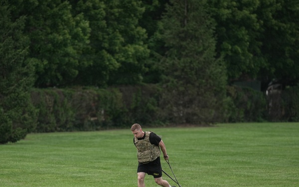 A male athlete is performing a resistance sled drag exercise on a well-maintained green grass field. He is wearing military-style workout attire including a camouflage plate carrier vest, black shorts with 'ARMY' text, and is pulling a weighted sled using straps. The setting appears to be an open field surrounded by dense green trees, suggesting an outdoor military or athletic training session.