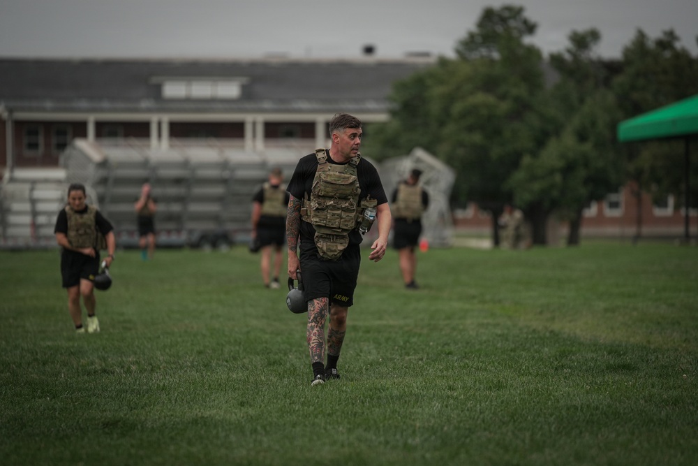 A group of individuals in military physical training attire are exercising on a grass field, wearing tactical plate carriers and carrying kettlebells. The foreground subject, wearing black shorts labeled 'ARMY' and a multicam plate carrier, has visible tattoos on his legs and arms. The setting appears to be a military installation based on the brick buildings and bleachers visible in the background.