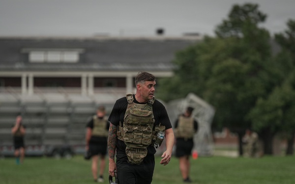A group of individuals in military physical training attire are exercising on a grass field, wearing tactical plate carriers and carrying kettlebells. The foreground subject, wearing black shorts labeled 'ARMY' and a multicam plate carrier, has visible tattoos on his legs and arms. The setting appears to be a military installation based on the brick buildings and bleachers visible in the background.