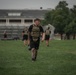 A group of individuals in military physical training attire are exercising on a grass field, wearing tactical plate carriers and carrying kettlebells. The foreground subject, wearing black shorts labeled 'ARMY' and a multicam plate carrier, has visible tattoos on his legs and arms. The setting appears to be a military installation based on the brick buildings and bleachers visible in the background.