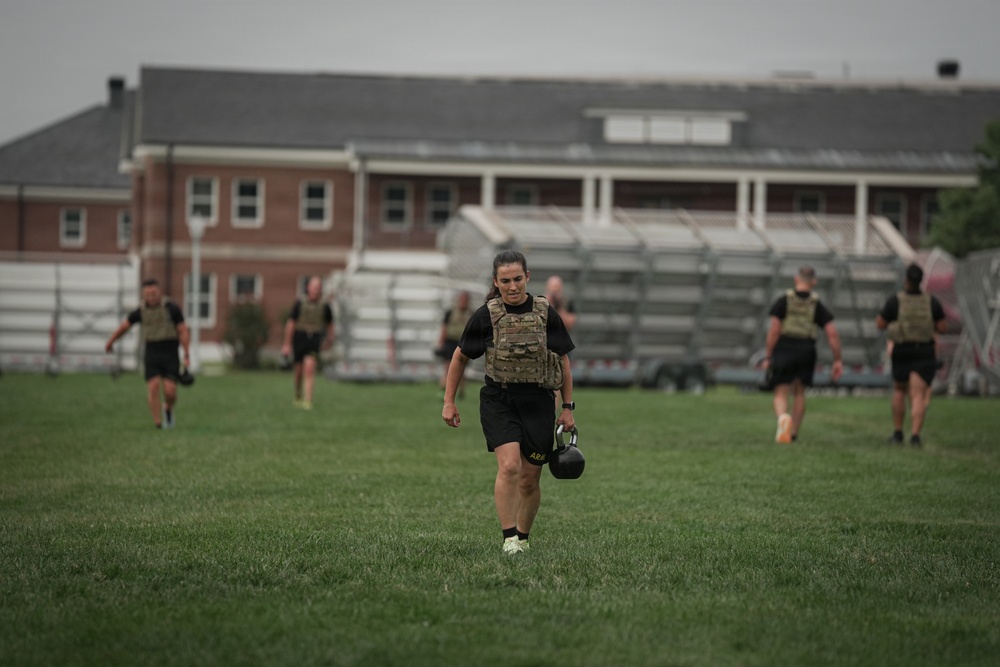 A group of military personnel are conducting physical training on a grass field, wearing black PT uniforms and tactical plate carriers/vests. The foreground features a female soldier carrying a kettlebell while walking toward the camera. A brick building with bleachers is visible in the background, suggesting a military installation or training facility.