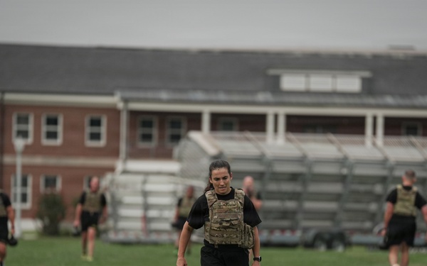 A group of military personnel are conducting physical training on a grass field, wearing black PT uniforms and tactical plate carriers/vests. The foreground features a female soldier carrying a kettlebell while walking toward the camera. A brick building with bleachers is visible in the background, suggesting a military installation or training facility.