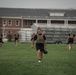 A group of military personnel are conducting physical training on a grass field, wearing black PT uniforms and tactical plate carriers/vests. The foreground features a female soldier carrying a kettlebell while walking toward the camera. A brick building with bleachers is visible in the background, suggesting a military installation or training facility.