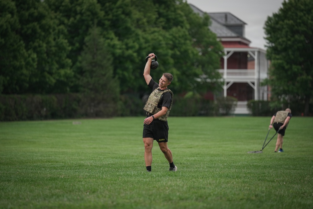 Two individuals are performing outdoor physical training exercises on a well-maintained grass field. The foreground subject, wearing Army-branded shorts and a tactical/plate carrier vest, is performing a single-arm kettlebell overhead press or swing. A second person is visible in the background, bent over and pulling a resistance sled or rope.