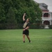 Two individuals are performing outdoor physical training exercises on a well-maintained grass field. The foreground subject, wearing Army-branded shorts and a tactical/plate carrier vest, is performing a single-arm kettlebell overhead press or swing. A second person is visible in the background, bent over and pulling a resistance sled or rope.
