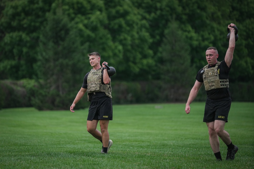 Two U.S. Army soldiers are conducting physical training on a grass field, each carrying a kettlebell. Both are wearing black Army PT shorts, black t-shirts, and tactical plate carriers/weight vests. One soldier carries the kettlebell at shoulder level while the other holds it overhead with one arm extended.