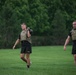 Two U.S. Army soldiers are conducting physical training on a grass field, each carrying a kettlebell. Both are wearing black Army PT shorts, black t-shirts, and tactical plate carriers/weight vests. One soldier carries the kettlebell at shoulder level while the other holds it overhead with one arm extended.