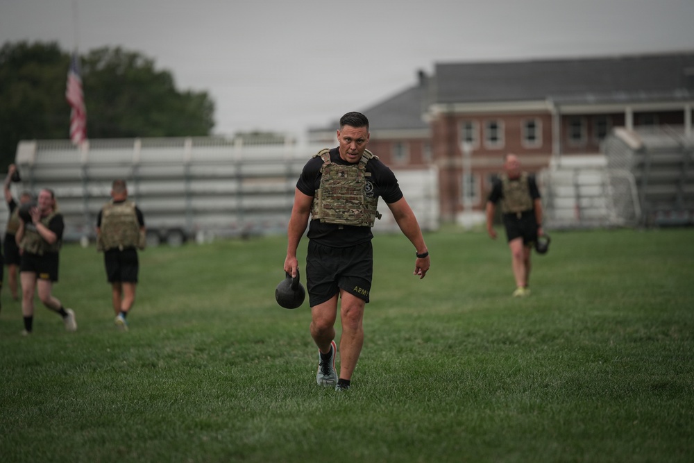 A group of individuals wearing black Army PT uniforms and tactical plate carriers are conducting a physical training exercise on a grass field. The primary subject in the foreground is carrying a kettlebell while walking purposefully across the field. An American flag and brick military-style buildings are visible in the background, suggesting a military installation setting.