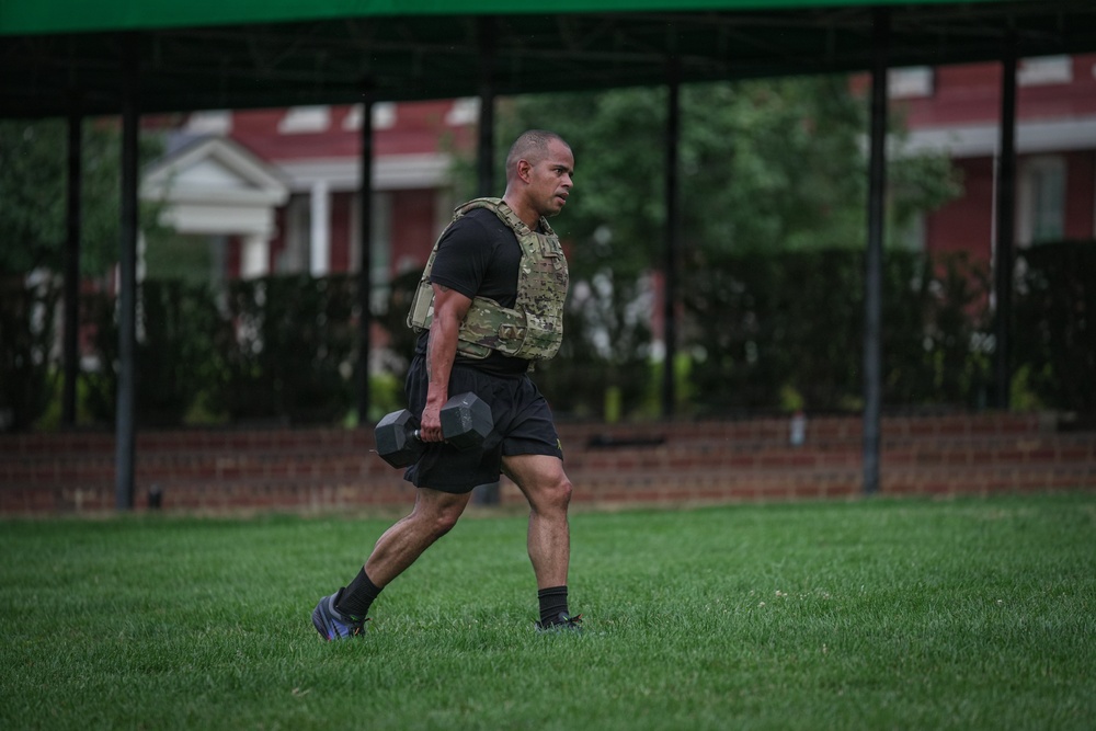 A muscular man is walking across a grass field while carrying a dumbbell in his right hand and wearing a camouflage tactical/weighted vest over a black t-shirt and black shorts. The setting appears to be an outdoor fitness training area, likely on a military installation based on the brick buildings and structured grounds visible in the background. The image captures a fitness or physical training exercise in progress.
