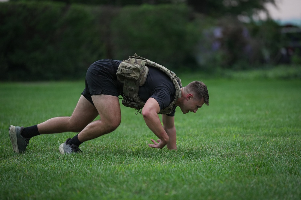 A young male is performing a bear crawl or ground movement exercise on a grass field while wearing a tactical/plate carrier vest over a black t-shirt and shorts. The exercise appears to be part of a military-style or tactical fitness training session. The setting is an outdoor grassy area with trees visible in the blurred background.