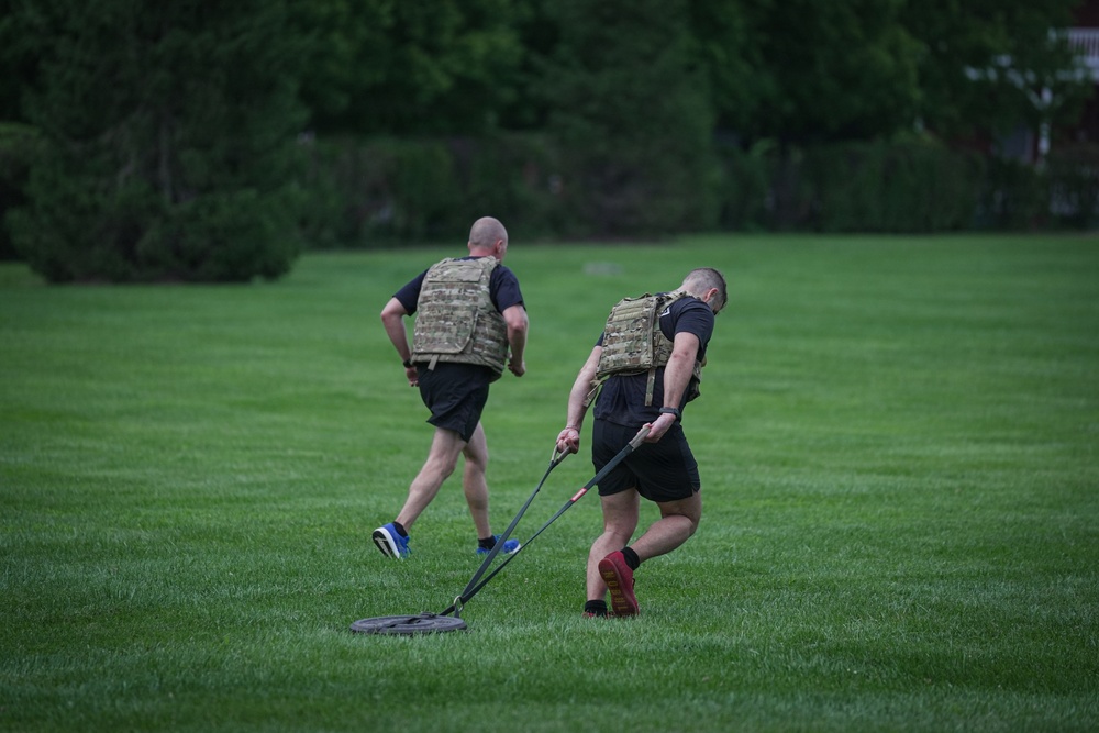Two individuals wearing tactical/plate carrier vests and black athletic clothing are performing an outdoor fitness workout on a well-maintained grass field. The person in the foreground is dragging a weighted sled using resistance straps, while the person in the background appears to be walking or running. The setting appears to be a park or athletic field with dense green trees in the background.