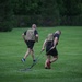 Two individuals wearing tactical/plate carrier vests and black athletic clothing are performing an outdoor fitness workout on a well-maintained grass field. The person in the foreground is dragging a weighted sled using resistance straps, while the person in the background appears to be walking or running. The setting appears to be a park or athletic field with dense green trees in the background.