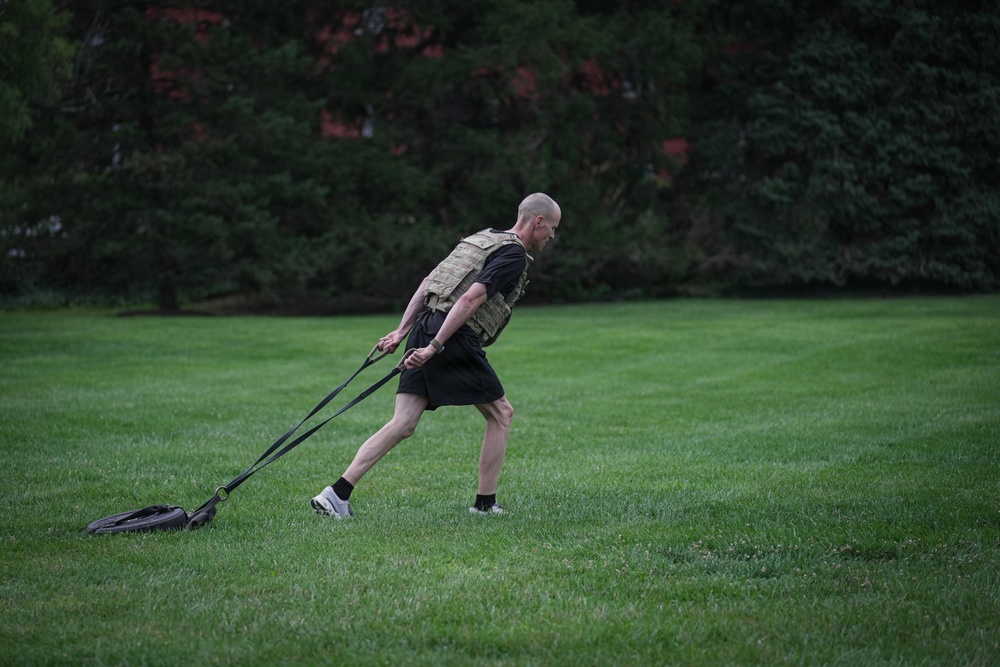 A bald man is performing an outdoor fitness workout on a well-maintained grass field. He is wearing a tactical/weighted vest over a black t-shirt and shorts, dragging a weighted sled using two straps. The setting appears to be a park or large lawn area with dense trees and a brick structure visible in the background.