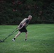 A bald man is performing an outdoor fitness workout on a well-maintained grass field. He is wearing a tactical/weighted vest over a black t-shirt and shorts, dragging a weighted sled using two straps. The setting appears to be a park or large lawn area with dense trees and a brick structure visible in the background.