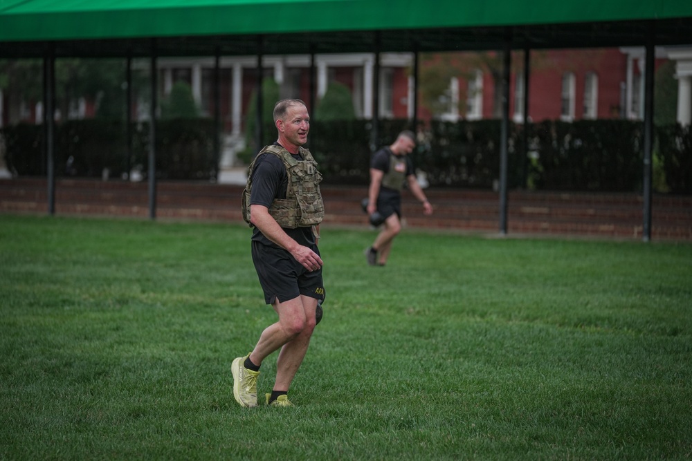 A middle-aged man wearing a tactical/weighted vest, black shorts, and neon yellow running shoes is walking or jogging on a well-maintained grass field. A second person in black athletic wear is visible in the background, also moving across the field. The setting appears to be a military installation or institutional campus, indicated by the brick buildings and green canopy tent structure.