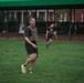 A middle-aged man wearing a tactical/weighted vest, black shorts, and neon yellow running shoes is walking or jogging on a well-maintained grass field. A second person in black athletic wear is visible in the background, also moving across the field. The setting appears to be a military installation or institutional campus, indicated by the brick buildings and green canopy tent structure.