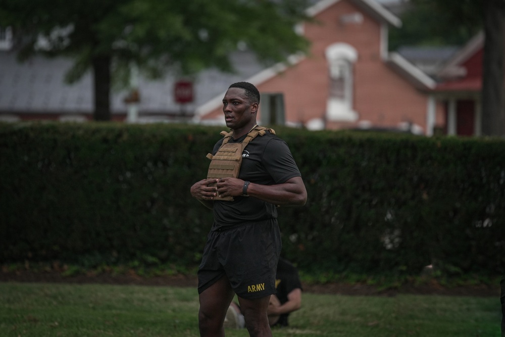 A male soldier wearing U.S. Army-branded black shorts and a tan plate carrier vest is engaged in an outdoor physical training exercise on a grassy area. He appears to be in motion, holding his hands together at chest level, possibly during a fitness drill or workout. The background features a trimmed hedge, trees, and a brick building, suggesting a military installation or base.