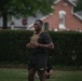 A male soldier wearing U.S. Army-branded black shorts and a tan plate carrier vest is engaged in an outdoor physical training exercise on a grassy area. He appears to be in motion, holding his hands together at chest level, possibly during a fitness drill or workout. The background features a trimmed hedge, trees, and a brick building, suggesting a military installation or base.