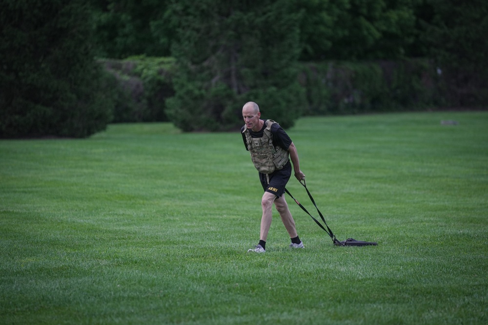 A bald male individual wearing military-style workout gear is performing a resistance sled drag exercise on a well-maintained grass field. He is wearing a tactical/plate carrier vest over a black shirt, black shorts with 'ARMY' printed in gold letters, and is pulling a weighted sled via straps. The background features dense green trees, suggesting an outdoor training environment.