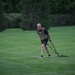 A bald male individual wearing military-style workout gear is performing a resistance sled drag exercise on a well-maintained grass field. He is wearing a tactical/plate carrier vest over a black shirt, black shorts with 'ARMY' printed in gold letters, and is pulling a weighted sled via straps. The background features dense green trees, suggesting an outdoor training environment.