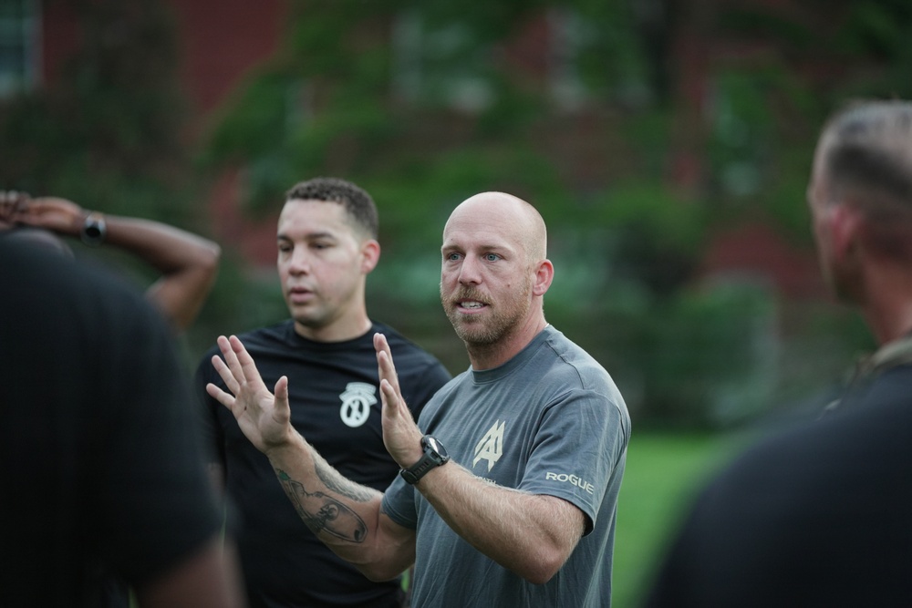 A bald man wearing a gray 'Rogue' branded t-shirt with visible arm tattoos is speaking and gesturing with both hands raised, apparently instructing or briefing a group outdoors. Behind him stands another man in a black t-shirt with a logo emblem. The setting appears to be a grassy area near a red brick building.