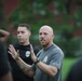 A bald man wearing a gray 'Rogue' branded t-shirt with visible arm tattoos is speaking and gesturing with both hands raised, apparently instructing or briefing a group outdoors. Behind him stands another man in a black t-shirt with a logo emblem. The setting appears to be a grassy area near a red brick building.