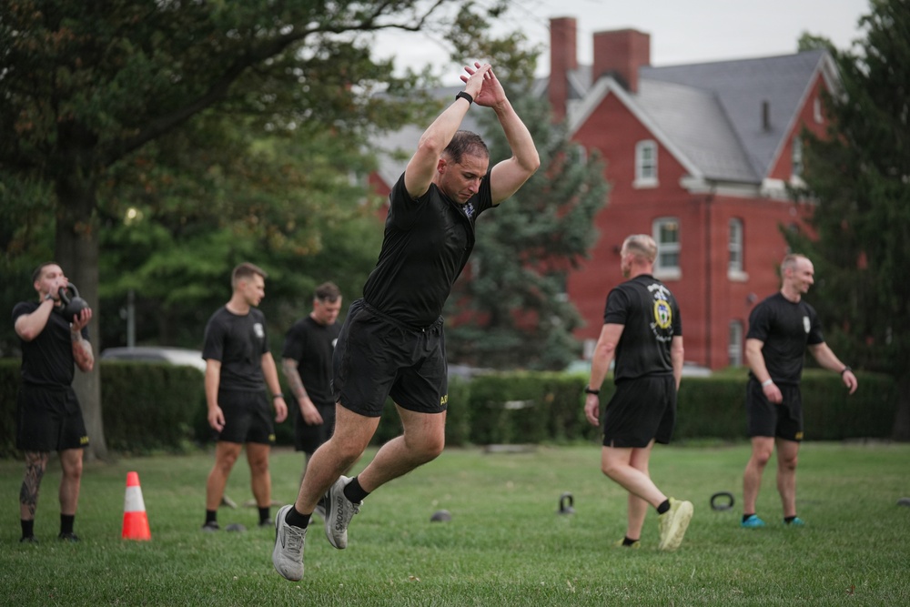 A group of individuals dressed in matching black athletic uniforms are conducting an outdoor physical training session on a grass field. The foreground subject is mid-jump with arms raised overhead, while others in the background perform various exercises including kettlebell work. The setting appears to be a military or institutional campus, evidenced by the red brick buildings and manicured grounds in the background.