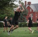 A group of individuals dressed in matching black athletic uniforms are conducting an outdoor physical training session on a grass field. The foreground subject is mid-jump with arms raised overhead, while others in the background perform various exercises including kettlebell work. The setting appears to be a military or institutional campus, evidenced by the red brick buildings and manicured grounds in the background.