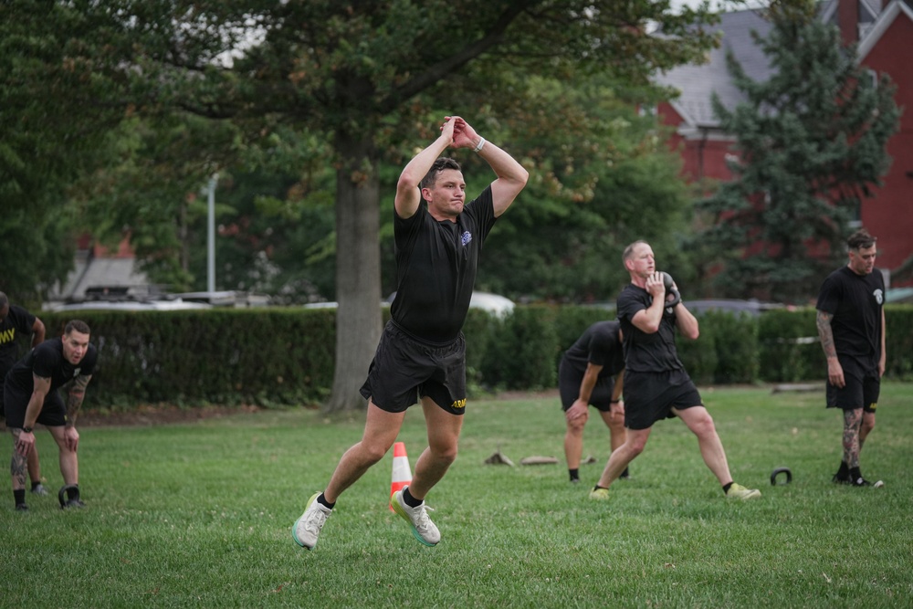A group of U.S. Army soldiers in black PT (physical training) uniforms are conducting an outdoor fitness workout on a grass field. The foreground soldier appears to be performing a kettlebell swing or similar explosive movement with arms raised overhead. The setting appears to be a military installation or park with red brick buildings and trees in the background.