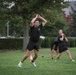 A group of U.S. Army soldiers in black PT (physical training) uniforms are conducting an outdoor fitness workout on a grass field. The foreground soldier appears to be performing a kettlebell swing or similar explosive movement with arms raised overhead. The setting appears to be a military installation or park with red brick buildings and trees in the background.