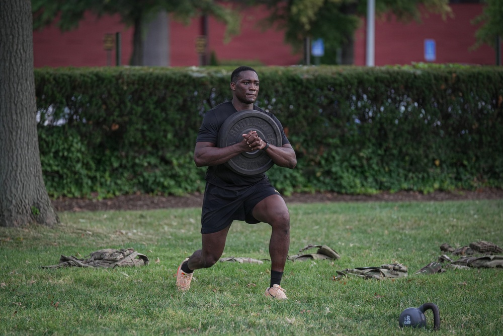 A muscular man is performing a weighted lunge exercise outdoors on a grass field, holding a barbell weight plate against his chest. He is dressed in all-black athletic wear with tan/orange sneakers and black ankle socks. The setting appears to be a park or institutional grounds with a red brick building and manicured hedges visible in the background.
