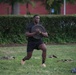 A muscular man is performing a weighted lunge exercise outdoors on a grass field, holding a barbell weight plate against his chest. He is dressed in all-black athletic wear with tan/orange sneakers and black ankle socks. The setting appears to be a park or institutional grounds with a red brick building and manicured hedges visible in the background.