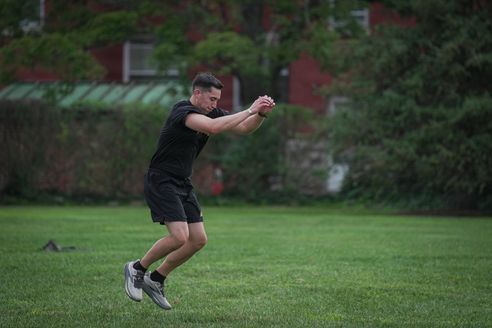 A young man dressed in all-black athletic wear is captured mid-air while performing what appears to be a broad jump or plyometric exercise on a grass field. His arms are extended forward and clasped together, and both feet are off the ground, indicating an explosive jumping movement. The background features a red brick building partially obscured by dense green foliage.