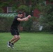 A young man dressed in all-black athletic wear is captured mid-air while performing what appears to be a broad jump or plyometric exercise on a grass field. His arms are extended forward and clasped together, and both feet are off the ground, indicating an explosive jumping movement. The background features a red brick building partially obscured by dense green foliage.