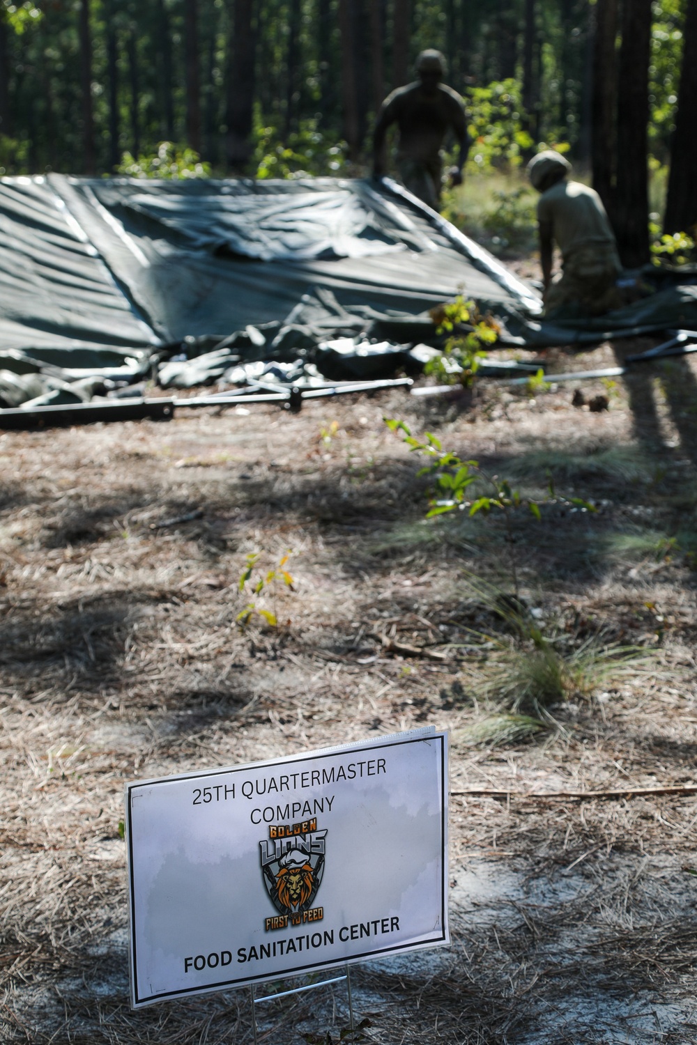A military field exercise showing personnel from the 25th Quartermaster Company setting up a Food Sanitation Center in a forested area. Two soldiers are visible in the background working to erect or dismantle a large dark military tent or tarpaulin structure among pine trees. A white sign in the foreground identifies the unit as the '25th Quartermaster Company - Golden Lions - First to Feed - Food Sanitation Center.'