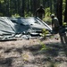 A military field exercise showing personnel from the 25th Quartermaster Company setting up a Food Sanitation Center in a forested area. Two soldiers are visible in the background working to erect or dismantle a large dark military tent or tarpaulin structure among pine trees. A white sign in the foreground identifies the unit as the '25th Quartermaster Company - Golden Lions - First to Feed - Food Sanitation Center.'