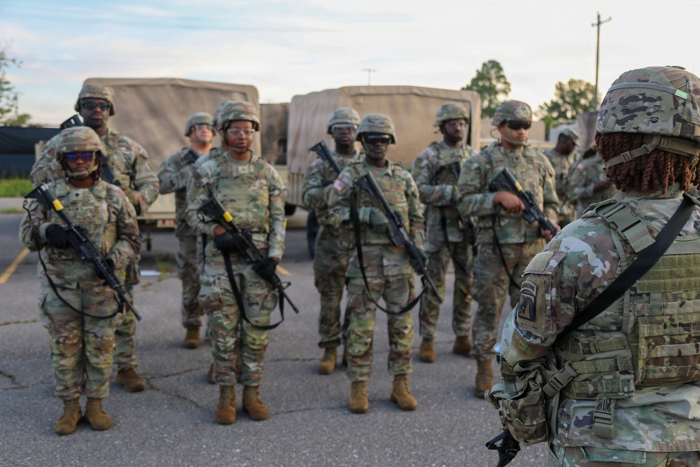 A group of approximately 8-9 soldiers in U.S. Army OCP (Operational Camouflage Pattern) uniforms are standing in formation on a paved surface. A soldier with their back to the camera, wearing an Airborne unit patch, appears to be addressing or briefing the assembled troops. Military vehicles, likely HMMWVs or similar tactical trucks, are visible in the background.