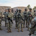 A group of approximately 8-9 soldiers in U.S. Army OCP (Operational Camouflage Pattern) uniforms are standing in formation on a paved surface. A soldier with their back to the camera, wearing an Airborne unit patch, appears to be addressing or briefing the assembled troops. Military vehicles, likely HMMWVs or similar tactical trucks, are visible in the background.