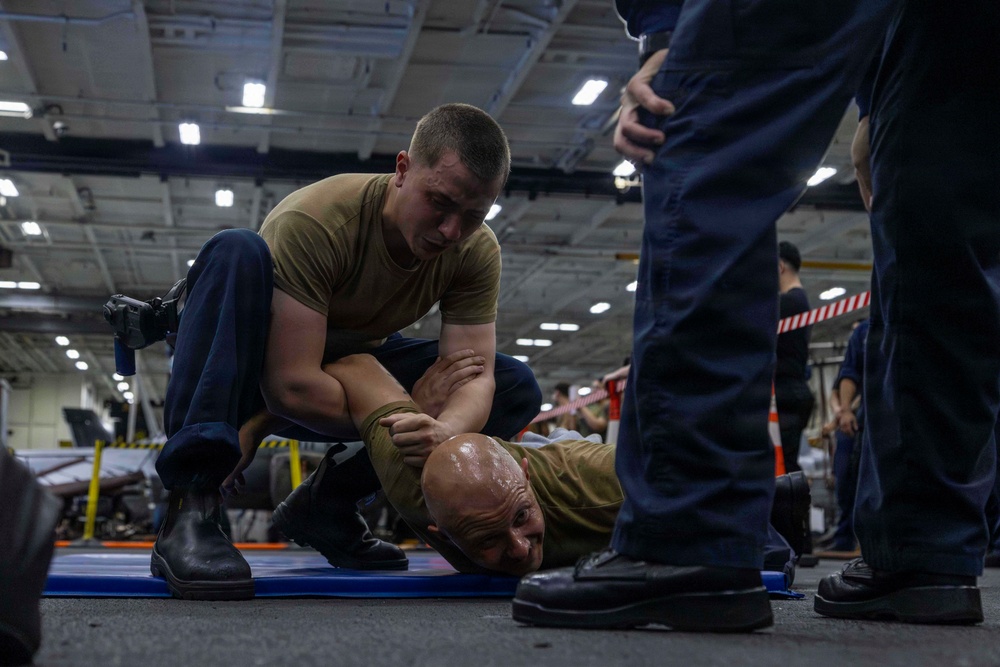 A military training exercise is taking place in what appears to be a ship's hangar bay or large indoor facility. One service member in a tan t-shirt is performing a ground control/restraint technique on another individual lying face-down on a blue mat. Observers in dark uniforms are watching the demonstration.