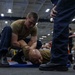 A military training exercise is taking place in what appears to be a ship's hangar bay or large indoor facility. One service member in a tan t-shirt is performing a ground control/restraint technique on another individual lying face-down on a blue mat. Observers in dark uniforms are watching the demonstration.