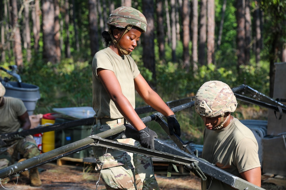 Two military personnel in camouflage uniforms and helmets are working together to assemble or disassemble a piece of military field equipment, likely a cot frame or folding support structure. The setting appears to be an outdoor training area or field camp surrounded by pine trees. A third soldier is partially visible in the background near additional equipment and supplies.