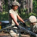 Two military personnel in camouflage uniforms and helmets are working together to assemble or disassemble a piece of military field equipment, likely a cot frame or folding support structure. The setting appears to be an outdoor training area or field camp surrounded by pine trees. A third soldier is partially visible in the background near additional equipment and supplies.