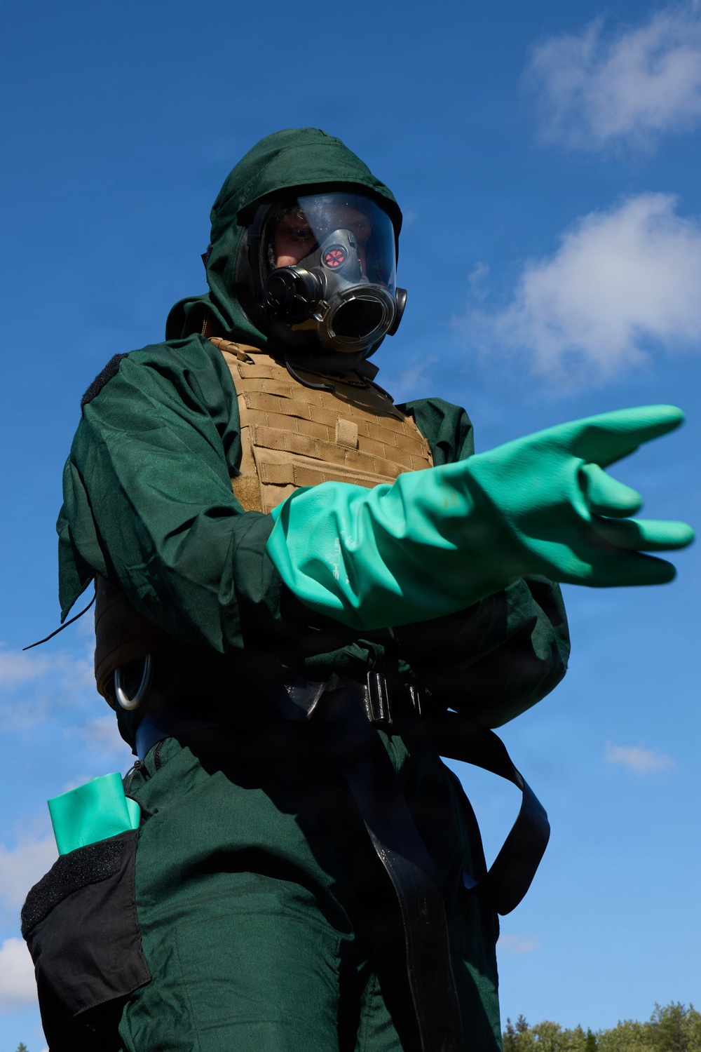 A person is wearing full CBRN (Chemical, Biological, Radiological, Nuclear) protective gear, including a green hazmat suit, full-face gas mask, and long green rubber gloves. They are also wearing a tan/coyote tactical plate carrier vest over the suit. The individual is extending one arm outward, possibly gesturing or directing, photographed from a low angle against a blue sky.