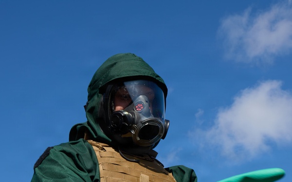 A person is wearing full CBRN (Chemical, Biological, Radiological, Nuclear) protective gear, including a green hazmat suit, full-face gas mask, and long green rubber gloves. They are also wearing a tan/coyote tactical plate carrier vest over the suit. The individual is extending one arm outward, possibly gesturing or directing, photographed from a low angle against a blue sky.