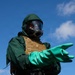 A person is wearing full CBRN (Chemical, Biological, Radiological, Nuclear) protective gear, including a green hazmat suit, full-face gas mask, and long green rubber gloves. They are also wearing a tan/coyote tactical plate carrier vest over the suit. The individual is extending one arm outward, possibly gesturing or directing, photographed from a low angle against a blue sky.