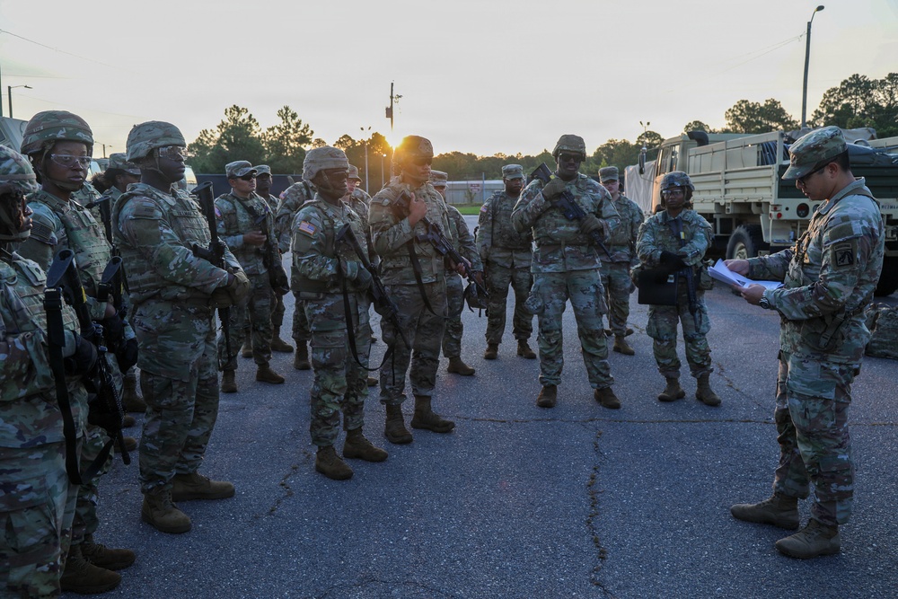 A group of U.S. Army soldiers in Operational Camouflage Pattern (OCP) uniforms are assembled in a parking area during early morning or evening hours, receiving a briefing from what appears to be a leader or NCO reading from documents. The soldiers are equipped with weapons, body armor, and tactical gear, suggesting a pre-mission or formation briefing. Military vehicles, including a medium tactical truck, are visible in the background.