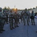 A group of U.S. Army soldiers in Operational Camouflage Pattern (OCP) uniforms are assembled in a parking area during early morning or evening hours, receiving a briefing from what appears to be a leader or NCO reading from documents. The soldiers are equipped with weapons, body armor, and tactical gear, suggesting a pre-mission or formation briefing. Military vehicles, including a medium tactical truck, are visible in the background.