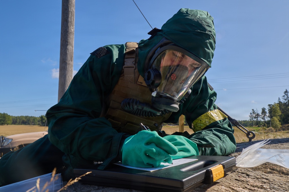 A person wearing full CBRN (Chemical, Biological, Radiological, Nuclear) protective gear, including a green hazmat suit, full-face gas mask, and green gloves, is writing on a document placed on a black case outdoors. The individual also wears a tan tactical vest over the protective suit. The setting appears to be a rural or field environment under clear blue skies, suggesting a military or emergency response exercise.