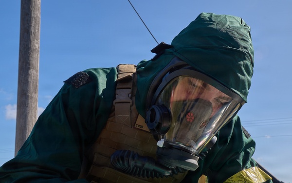 A person wearing full CBRN (Chemical, Biological, Radiological, Nuclear) protective gear, including a green hazmat suit, full-face gas mask, and green gloves, is writing on a document placed on a black case outdoors. The individual also wears a tan tactical vest over the protective suit. The setting appears to be a rural or field environment under clear blue skies, suggesting a military or emergency response exercise.